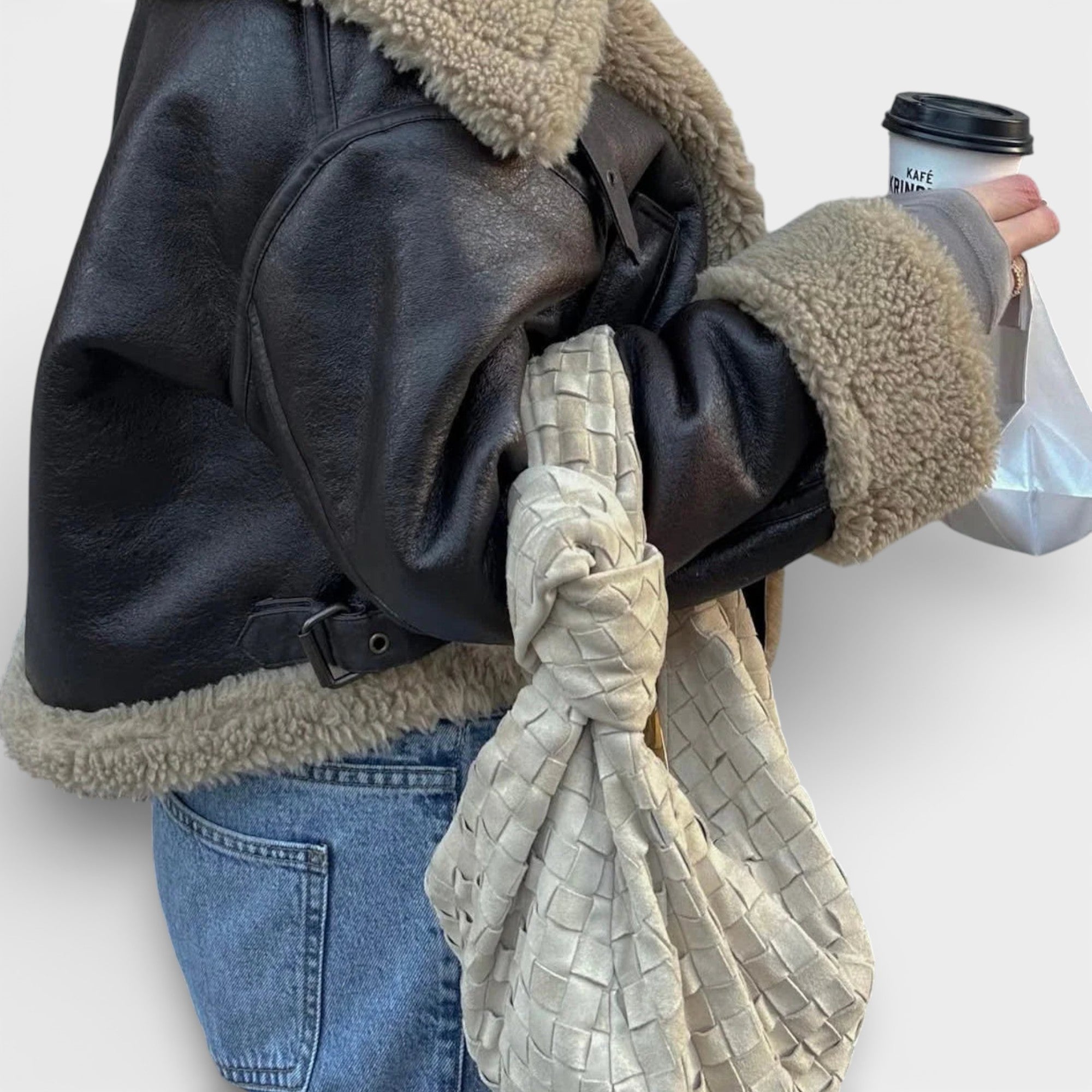 Person wearing a black leather jacket with shearling trim, holding a coffee cup and a woven bag.